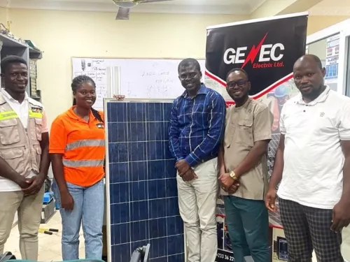 Cinco personas de pie en el interior alrededor de un gran panel solar, con una pancarta "GENEC Electrix Ltd." detr&aacute;s de ellos. El escenario es una oficina con dibujos t&eacute;cnicos en una pizarra al fondo.